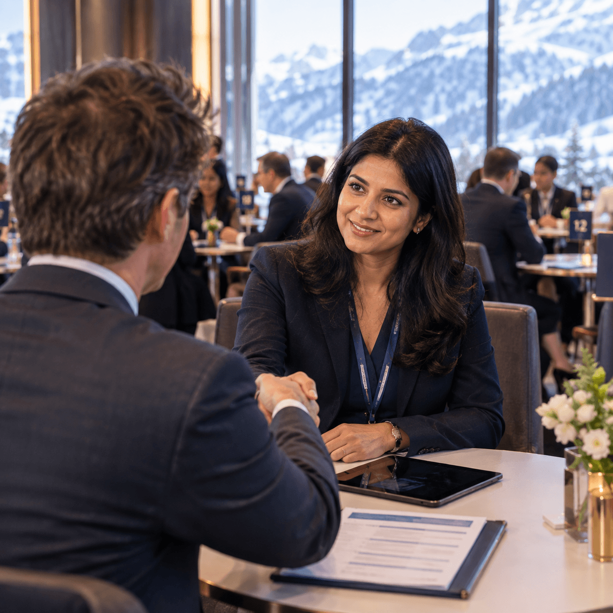 Two delegates shaking hands across a table at a one-to-one matchmaking meeting, other meetings and snowy mountains behind