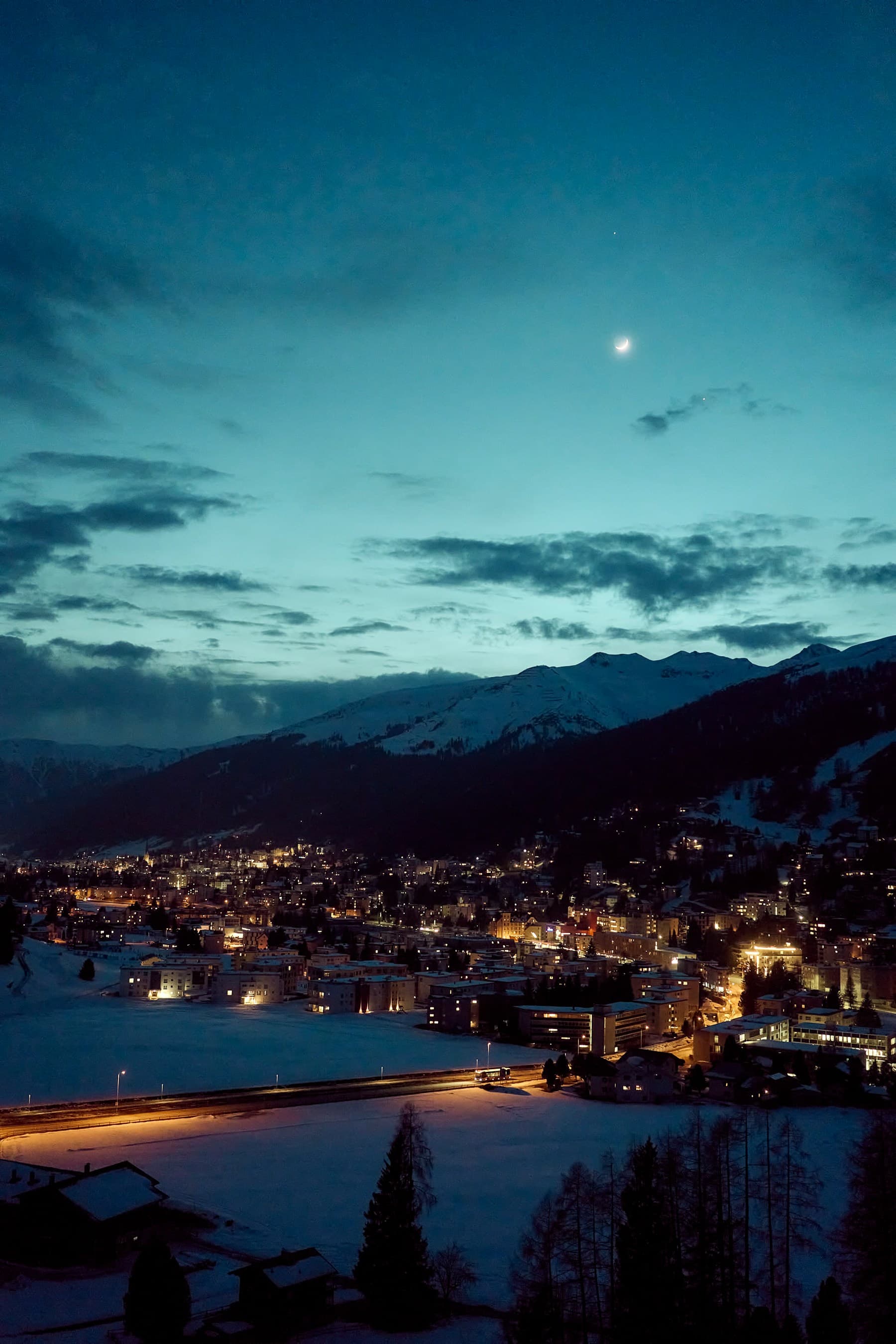 An Alpine town at dusk beneath snowy mountains and the moon
