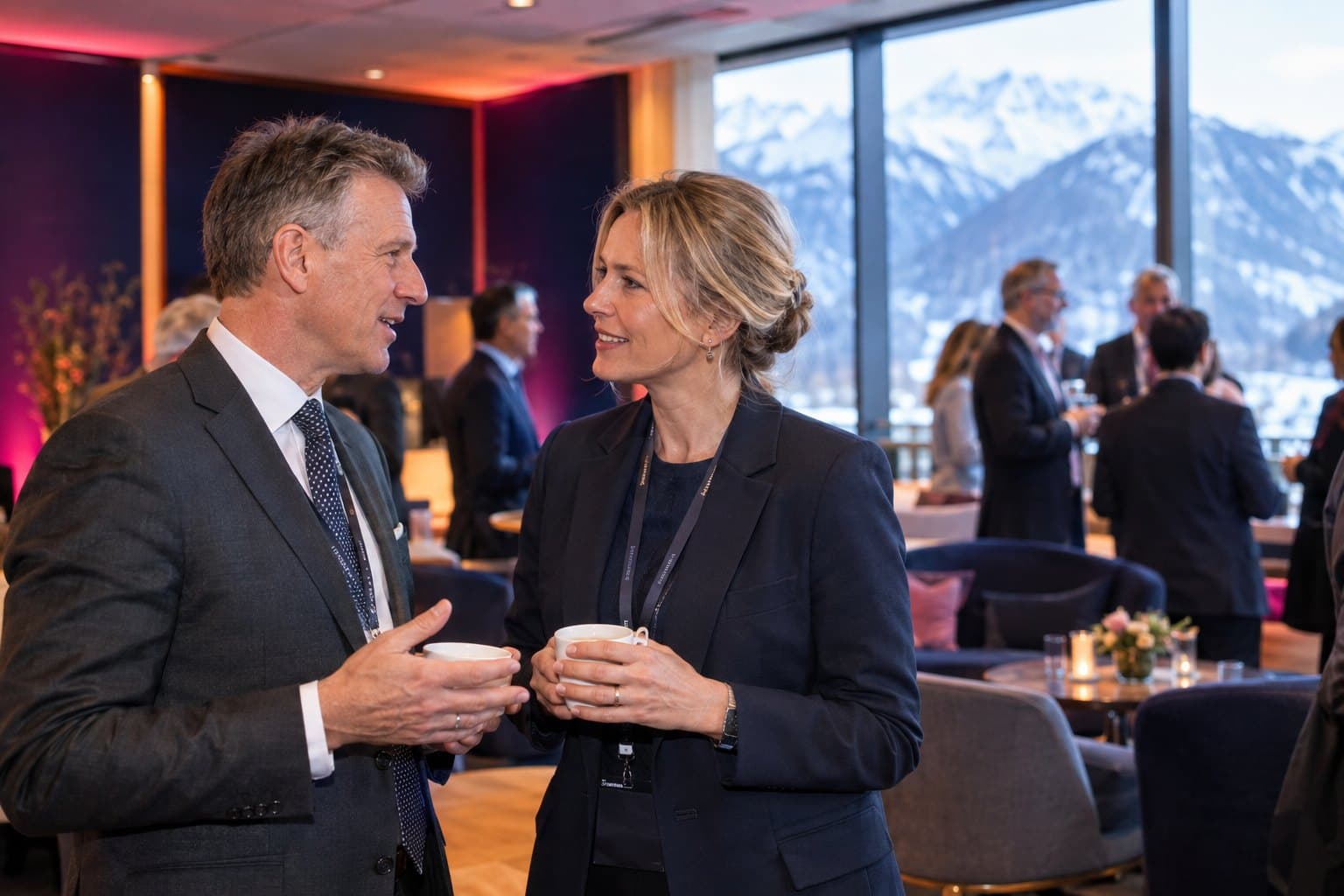 Two delegates in conversation over coffee in a summit lounge, with the snow-covered Alps through the windows